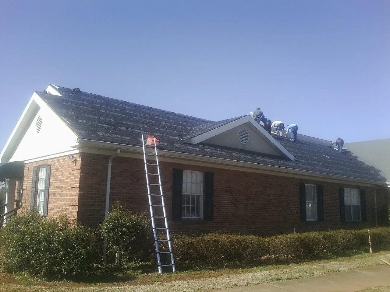 Crew tearing shingles off a brick ranch roof with a ladder set — Metal Roof Repair work by our crew in Log Lane Village