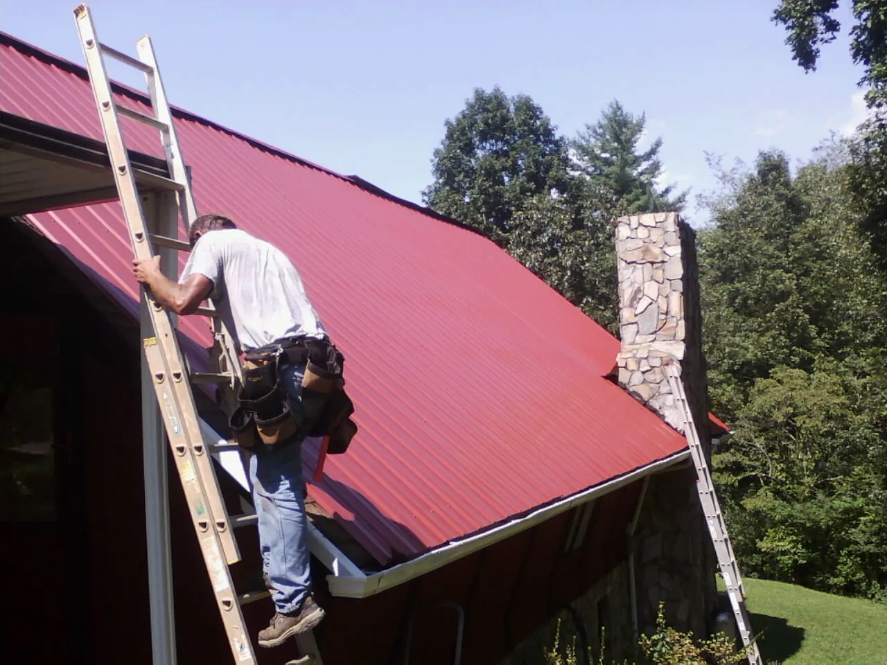 Roofer on a ladder beside a red metal roof and stone chimney — Soffit & Fascia Repair by Roofing Brothers in Log Lane Village