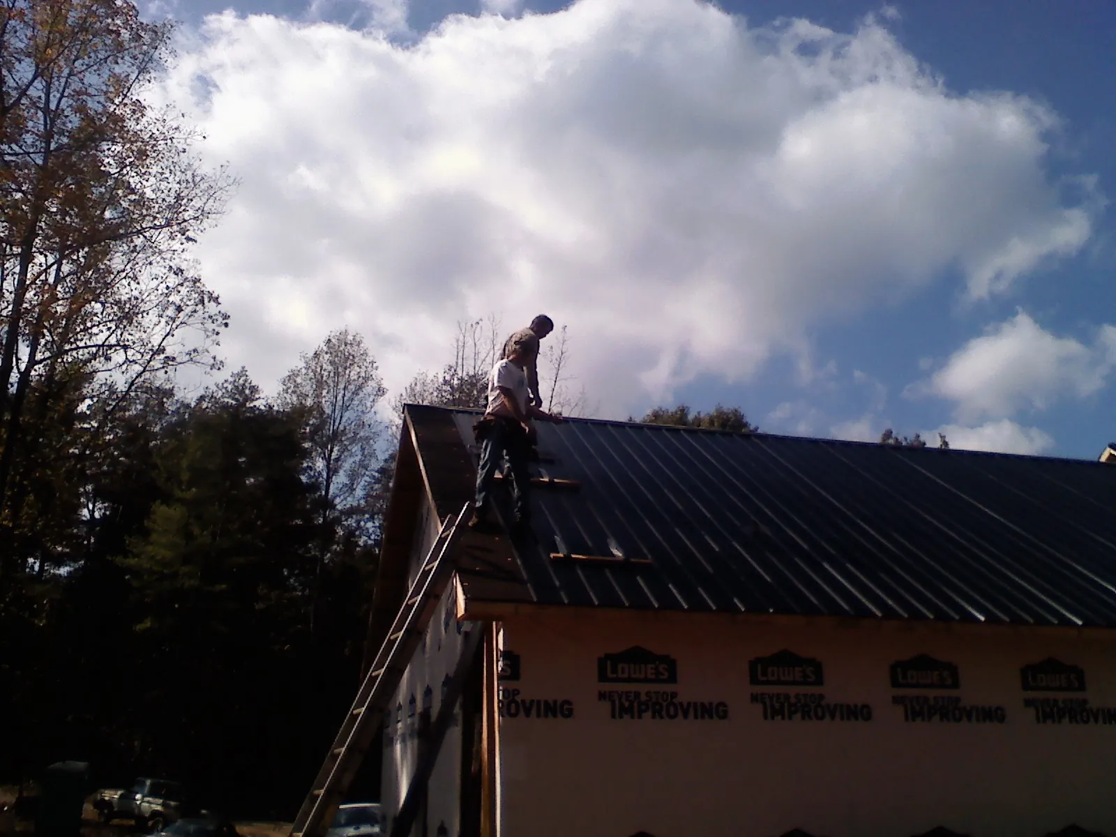 Roofer fastening dark metal panels high against a cloudy sky, from a Roof Inspection job in Log Lane Village